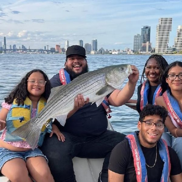 Group on a boat holding a large fish with a city skyline in the background.