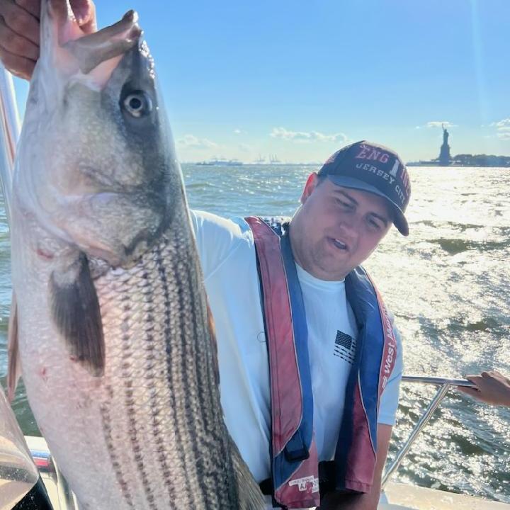 Person holding a large fish on a boat with water and the Statue of Liberty in the background.