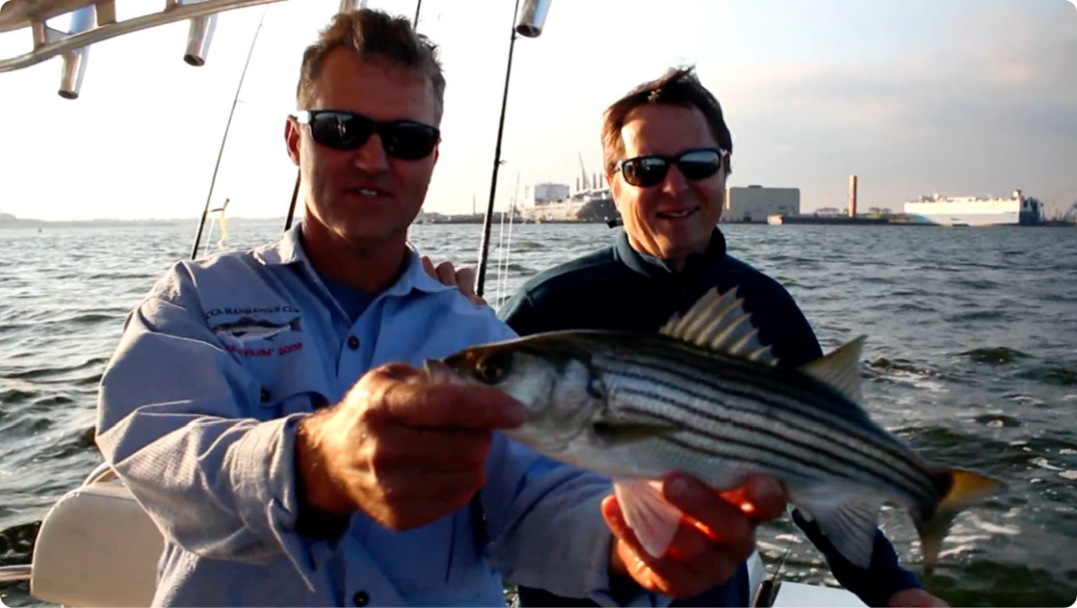 Two men on a boat holding a striped fish with water in the background.