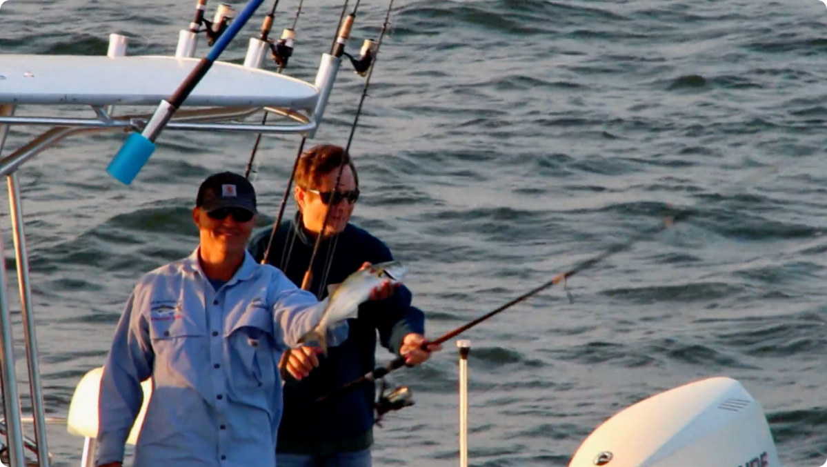 Two people fishing on a boat, one holds a caught fish.