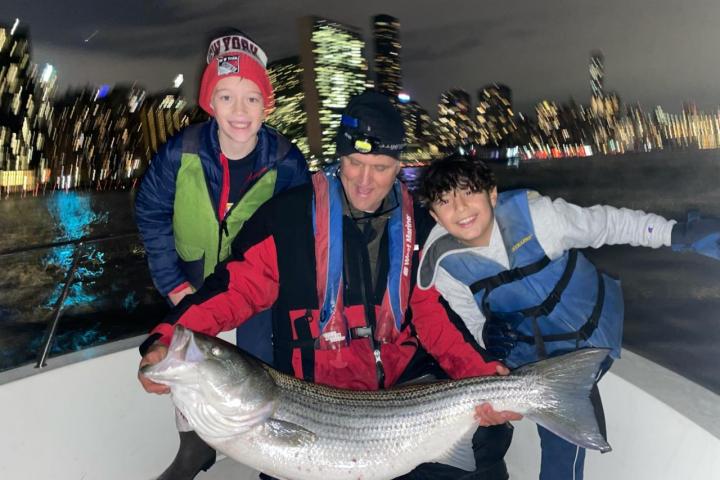 Three people on a boat at night holding a large fish with a city skyline in the background.