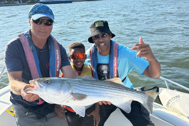 Three people on a boat holding a large fish with city skyline in background.