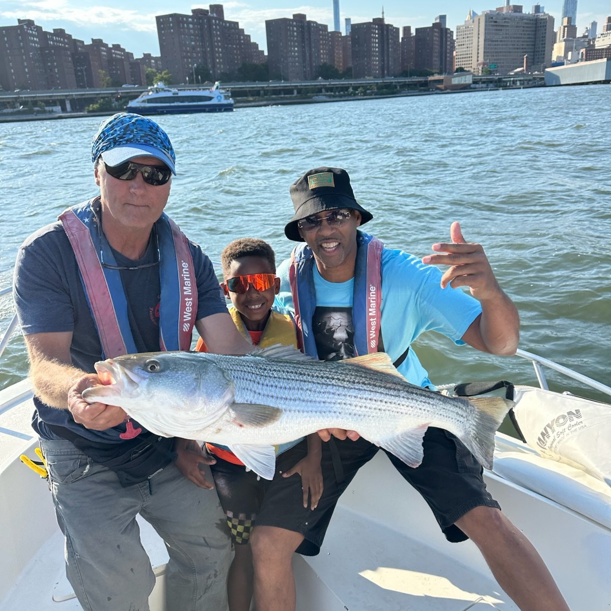 Three people on a boat holding a large fish with city skyline in background.