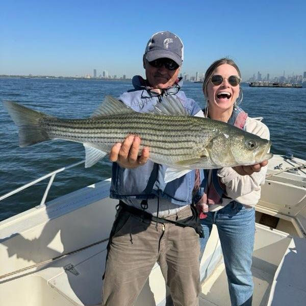 Two people on a boat holding a large fish, smiling against a blue sky.