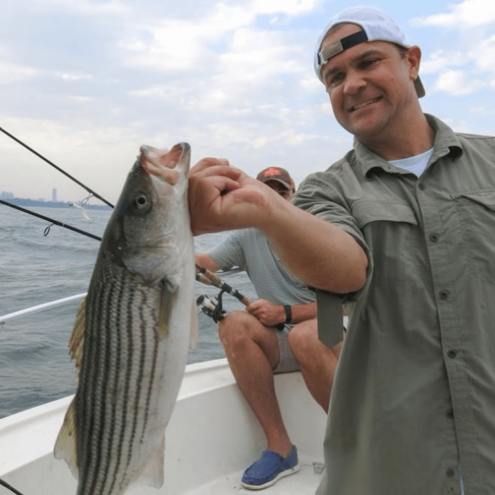 Man holding a striped bass fish on a boat with two fishing rods.