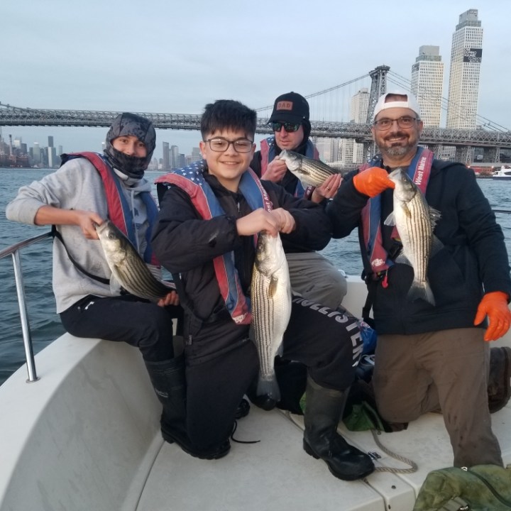 Four people on a boat holding fish with a city and bridge in the background.